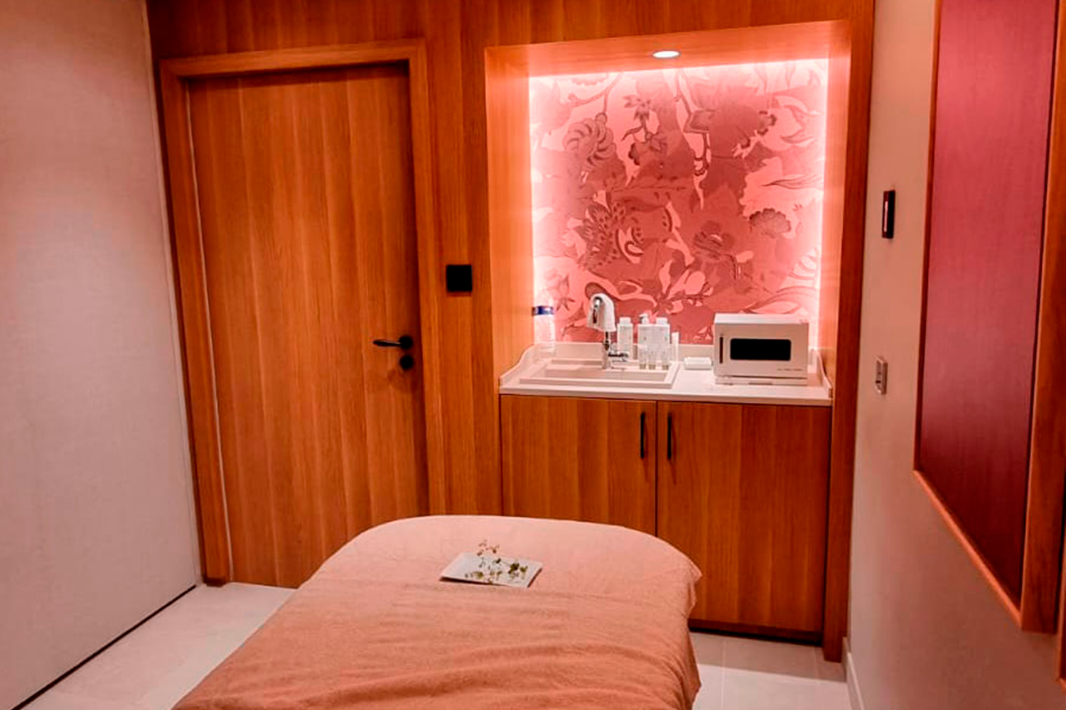 Un'accogliente sala per trattamenti spa con un lettino da massaggio coperto da un lenzuolo rosa, una porta e degli armadietti in legno, un bancone con prodotti da spa, un asciugamano e un microonde, il tutto illuminato da una calda luce rosa.