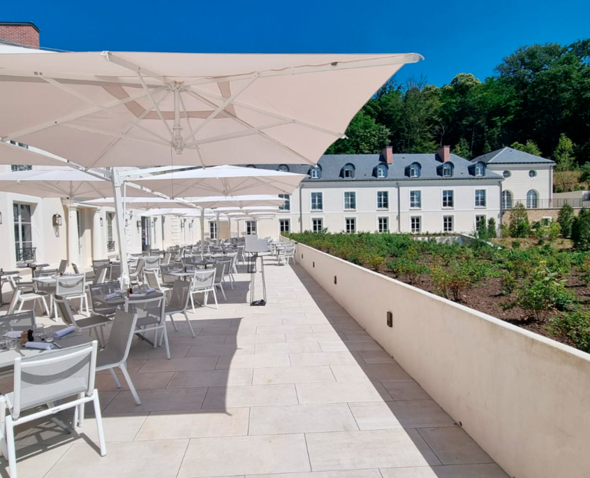 Un ampio patio esterno con tavoli bianchi, sedie e grandi ombrelloni si trova accanto a un imponente edificio bianco, sotto un cielo azzurro e con vista su un giardino con verde e alberi sullo sfondo.