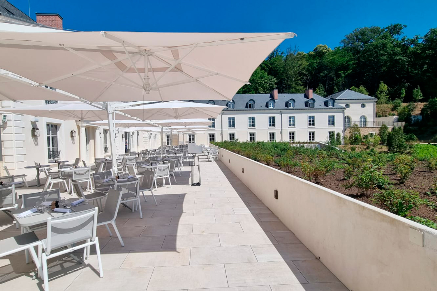 Un ampio patio esterno con tavoli bianchi, sedie e grandi ombrelloni si trova accanto a un imponente edificio bianco, sotto un cielo azzurro e con vista su un giardino con verde e alberi sullo sfondo.
