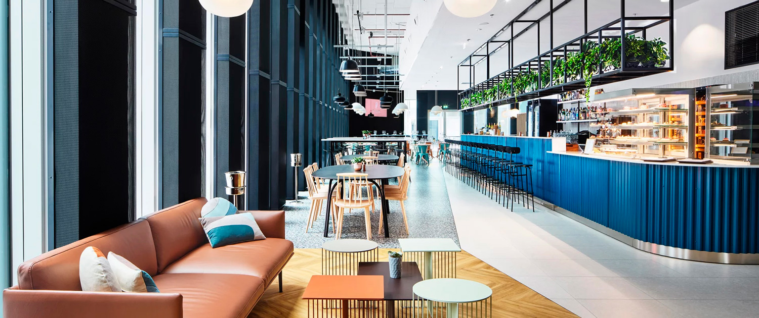 Moderna e luminosa zona soggiorno e pranzo di GH & Partners - Contract Division, con divano in pelle marrone, cuscini colorati, tavoli rotondi, un bancone blu con sgabelli alti, lampade a globo sospese e verde. Ampie finestre fiancheggiano il lato sinistro.