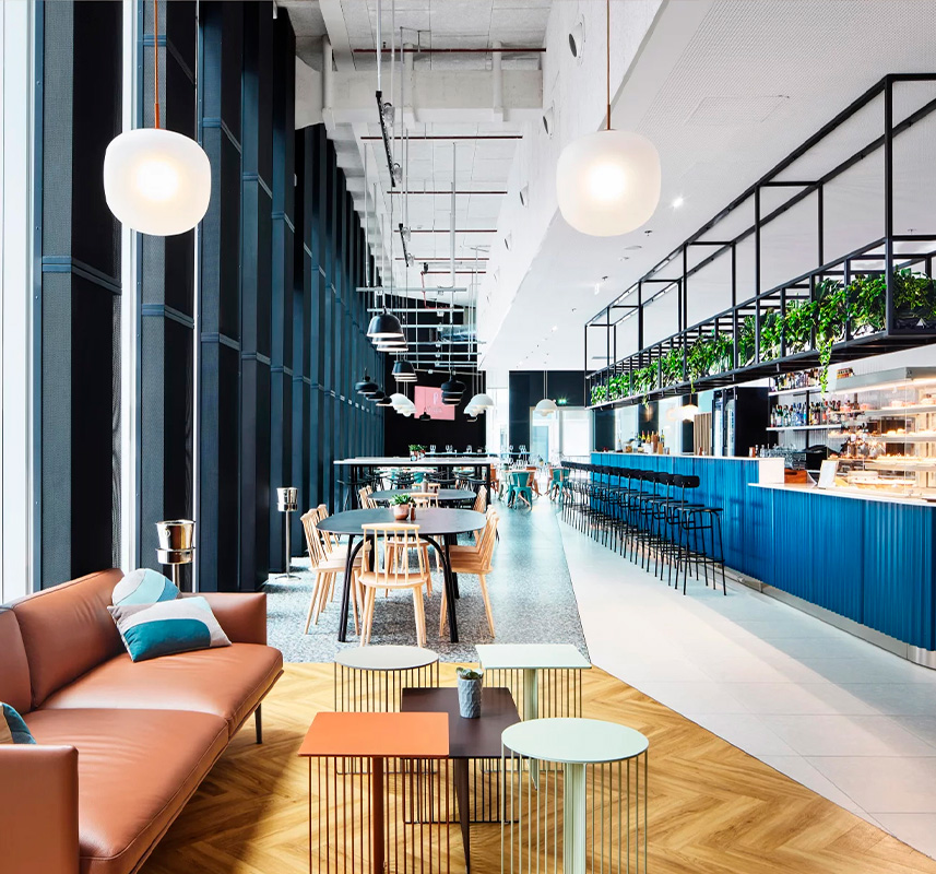 Caffetteria moderna e luminosa con ampie finestre, pareti con accenti blu e un divano in pelle marrone. Progettato da GH & Partners - Contract Division, questo spazio presenta progetti su misura: tavoli colorati, sedie in legno, un lungo bancone con sgabelli, lampade a globo e piante.