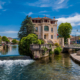 Un grande edificio con balconi si trova all'angolo di un piccolo canale con acqua limpida, circondato da alberi verdi, un ponte di pietra e posti a sedere all'aperto sotto un cielo blu con nuvole sparse.