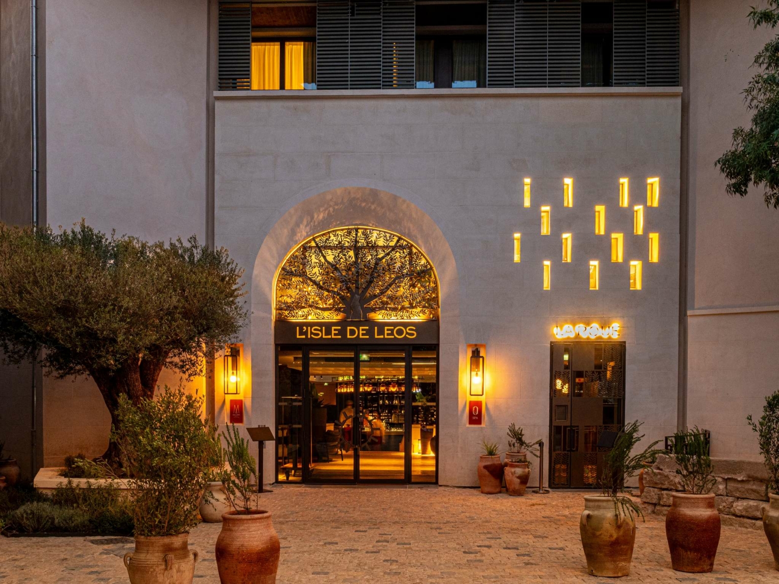 L'extérieur du restaurant L'Isle de Leos au crépuscule, niché près du Dolce by Wyndham Versailles, présente une grande entrée voûtée, des enseignes lumineuses, des appliques décoratives, des plantes en pot et un arbre dans une cour pavée.