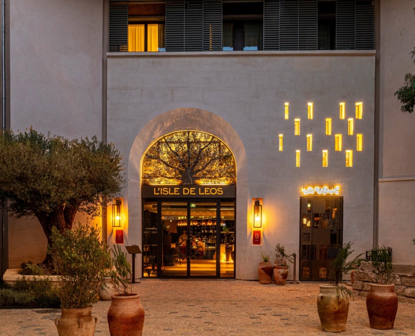 L'extérieur du restaurant L'Isle de Leos au crépuscule, niché près du Dolce by Wyndham Versailles, présente une grande entrée voûtée, des enseignes lumineuses, des appliques décoratives, des plantes en pot et un arbre dans une cour pavée.