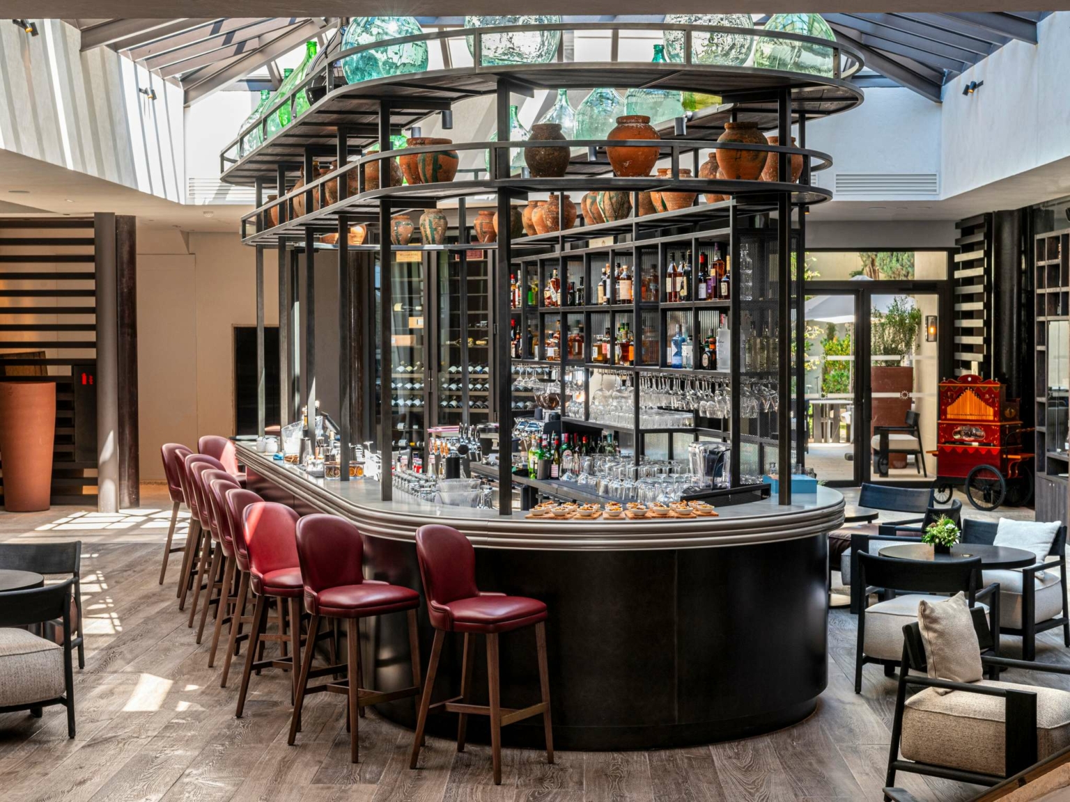 Bar intérieur moderne au Dolce by Wyndham Versailles | Domaine du Montcel, avec un comptoir incurvé, des tabourets de bar rouges, des étagères avec des verres, et la lumière naturelle qui se déverse à travers un plafond en verre. Des sièges confortables entourent ce bar accueillant.