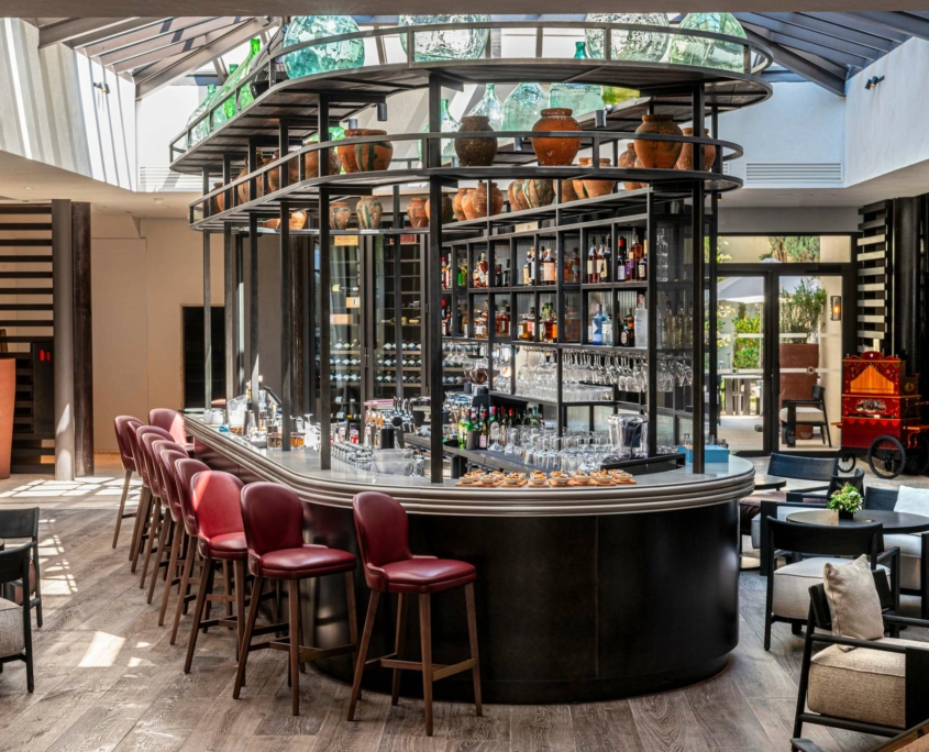 Bar intérieur moderne au Dolce by Wyndham Versailles | Domaine du Montcel, avec un comptoir incurvé, des tabourets de bar rouges, des étagères avec des verres, et la lumière naturelle qui se déverse à travers un plafond en verre. Des sièges confortables entourent ce bar accueillant.
