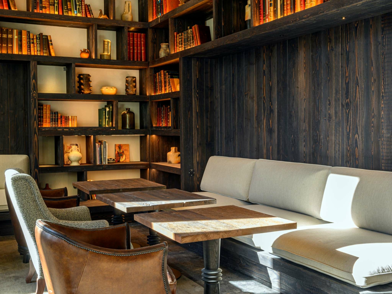 Coin salon confortable au Dolce by Wyndham Versailles, avec des bancs coussinés de couleur claire, des tables en bois et des fauteuils, entourés d'étagères en bois foncé remplies de livres et de décorations, le tout chaleureusement éclairé par la lumière naturelle au Domaine du Montcel.