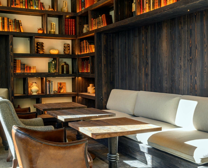 Coin salon confortable au Dolce by Wyndham Versailles, avec des bancs coussinés de couleur claire, des tables en bois et des fauteuils, entourés d'étagères en bois foncé remplies de livres et de décorations, le tout chaleureusement éclairé par la lumière naturelle au Domaine du Montcel.