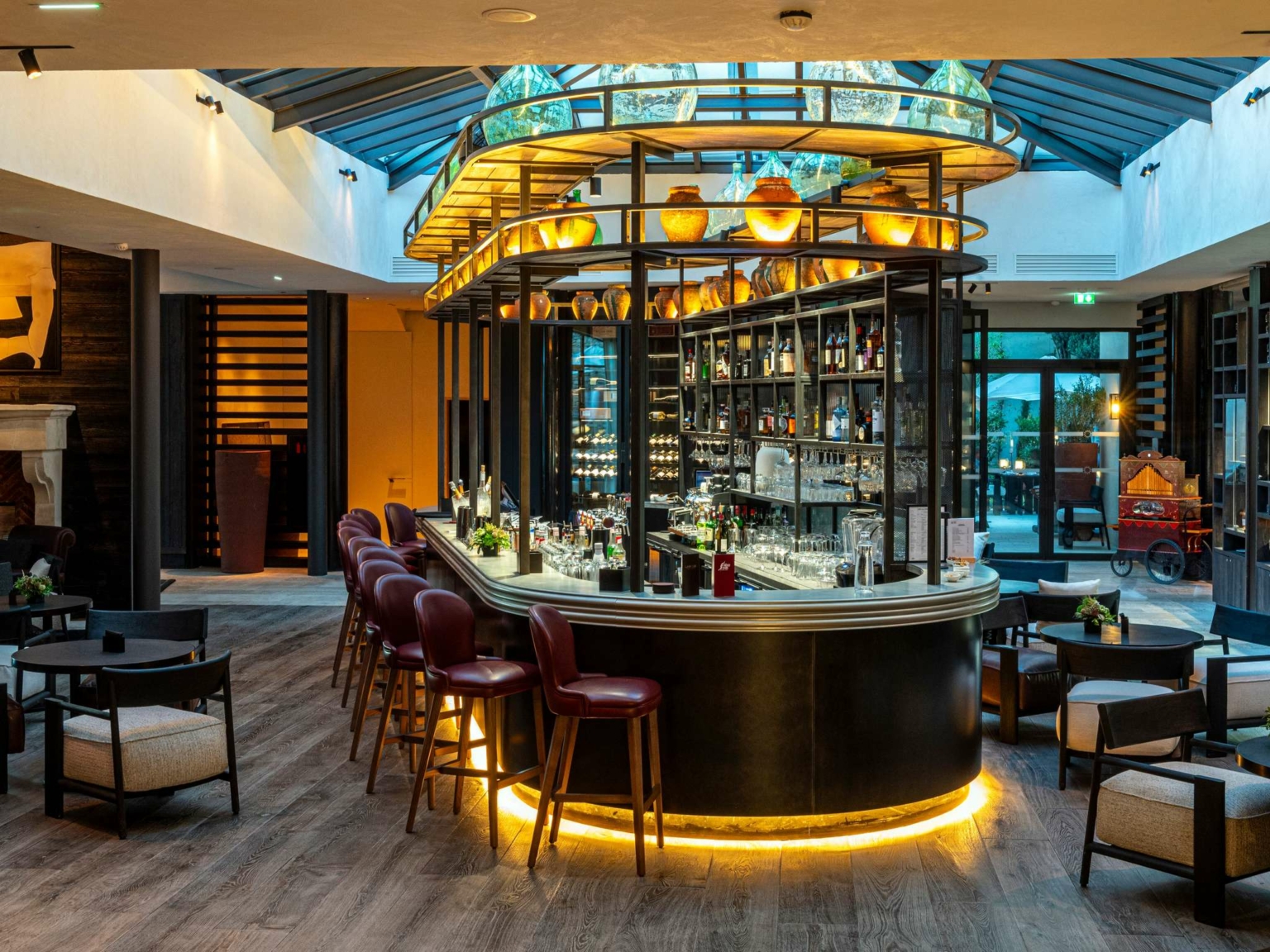 Bar moderne au Dolce by Wyndham Versailles avec un comptoir incurvé, des chaises à haut dossier et des étagères remplies de bouteilles. Un éclairage chaleureux, des planchers en bois et un plafond en verre créent une atmosphère accueillante au Domaine du Montcel.