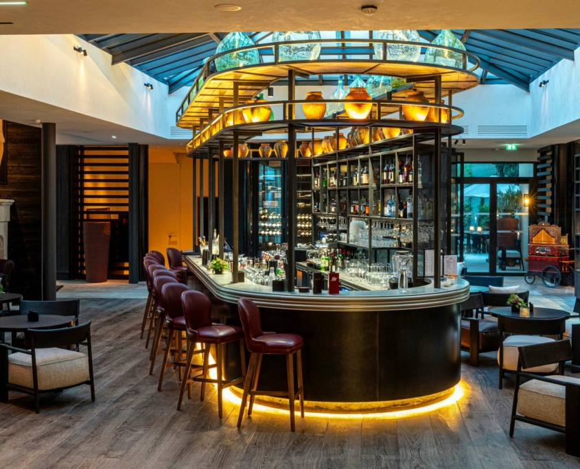 Bar moderne au Dolce by Wyndham Versailles avec un comptoir incurvé, des chaises à haut dossier et des étagères remplies de bouteilles. Un éclairage chaleureux, des planchers en bois et un plafond en verre créent une atmosphère accueillante au Domaine du Montcel.