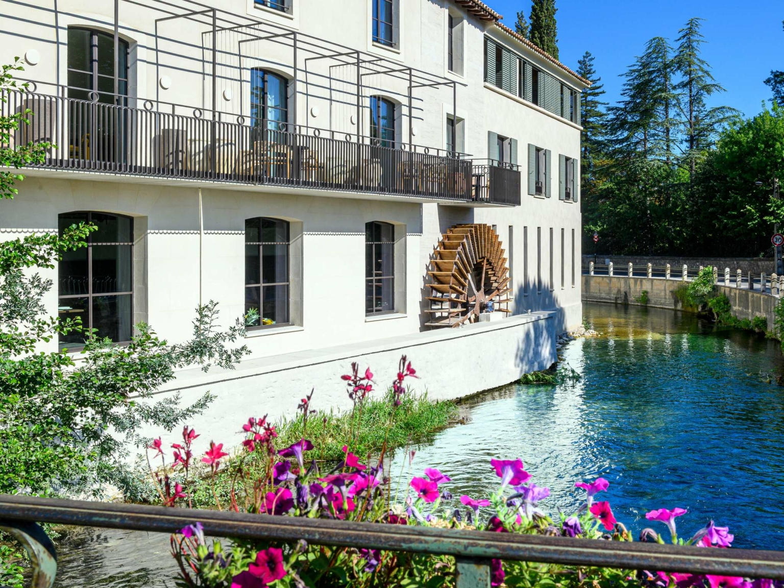 Un bâtiment blanc avec une roue à aubes en bois se dresse au bord d'une rivière limpide au Dolce by Wyndham Versailles | Domaine du Montcel. Des fleurs roses et violettes éclatantes s'épanouissent au premier plan, entourées d'arbres par une journée ensoleillée.