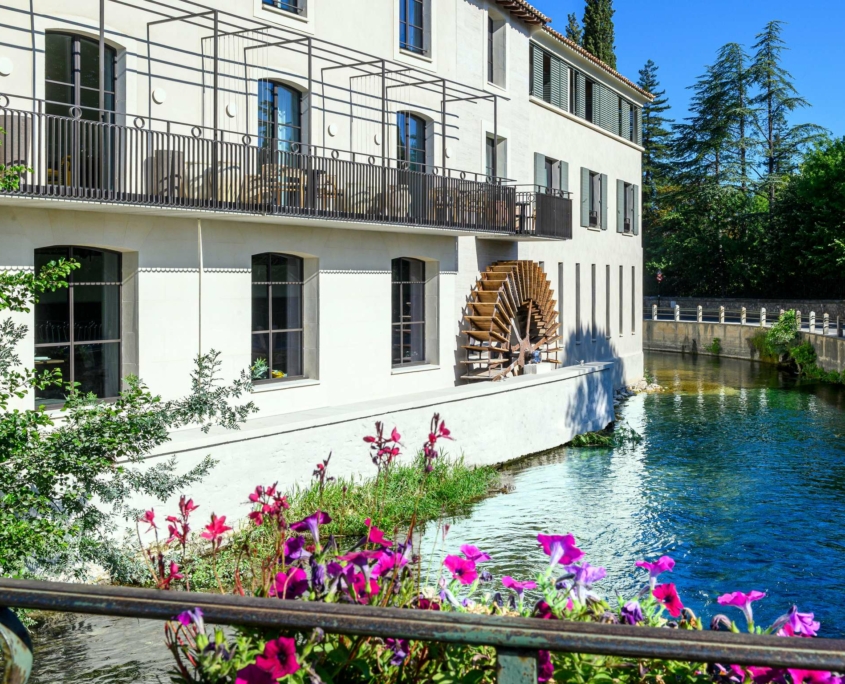 Un bâtiment blanc avec une roue à aubes en bois se dresse au bord d'une rivière limpide au Dolce by Wyndham Versailles | Domaine du Montcel. Des fleurs roses et violettes éclatantes s'épanouissent au premier plan, entourées d'arbres par une journée ensoleillée.