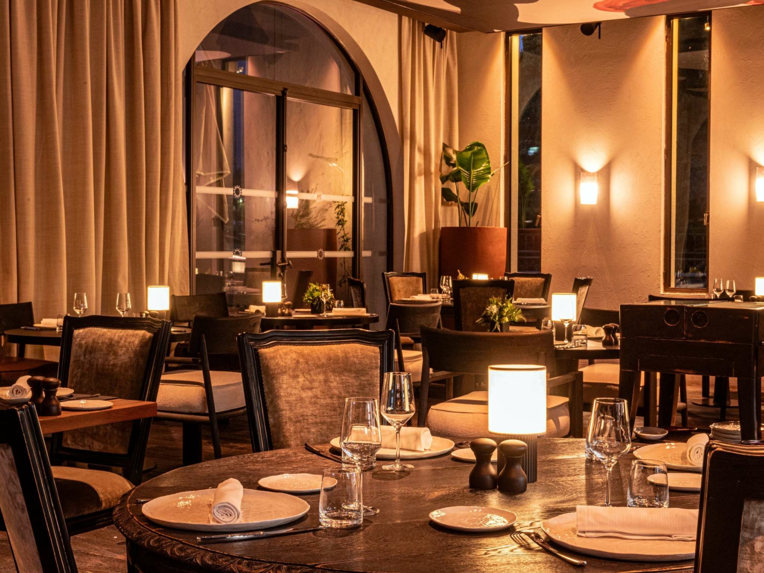 Intérieur élégant du restaurant Dolce by Wyndham Versailles avec des tables doucement éclairées, des chaises rembourrées, des serviettes blanches, de la verrerie et des plantes en pot. De grandes fenêtres et des appliques murales chaleureuses créent l'atmosphère chaleureuse et accueillante du Domaine du Montcel.