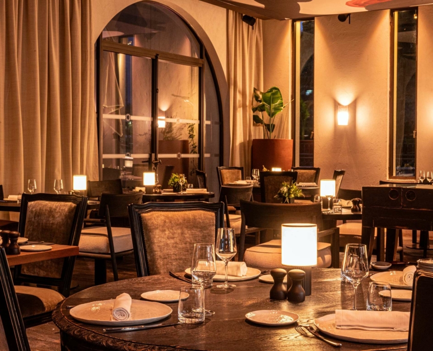 Intérieur élégant du restaurant Dolce by Wyndham Versailles avec des tables doucement éclairées, des chaises rembourrées, des serviettes blanches, de la verrerie et des plantes en pot. De grandes fenêtres et des appliques murales chaleureuses créent l'atmosphère chaleureuse et accueillante du Domaine du Montcel.