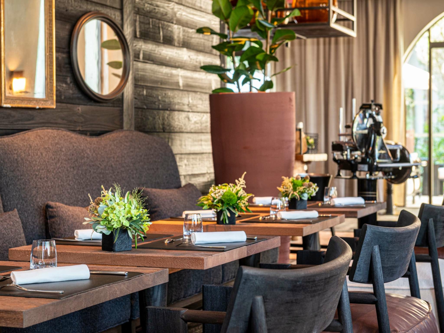 L'intérieur moderne du restaurant Dolce by Wyndham Versailles | Domaine du Montcel présente des tables en bois garnies de verres, de serviettes de table et de petits centres de table floraux. Des chaises sombres, une grande plante en pot et un éclairage doux complètent le décor neutre.