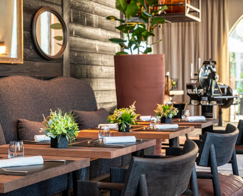 L'intérieur moderne du restaurant Dolce by Wyndham Versailles | Domaine du Montcel présente des tables en bois garnies de verres, de serviettes de table et de petits centres de table floraux. Des chaises sombres, une grande plante en pot et un éclairage doux complètent le décor neutre.