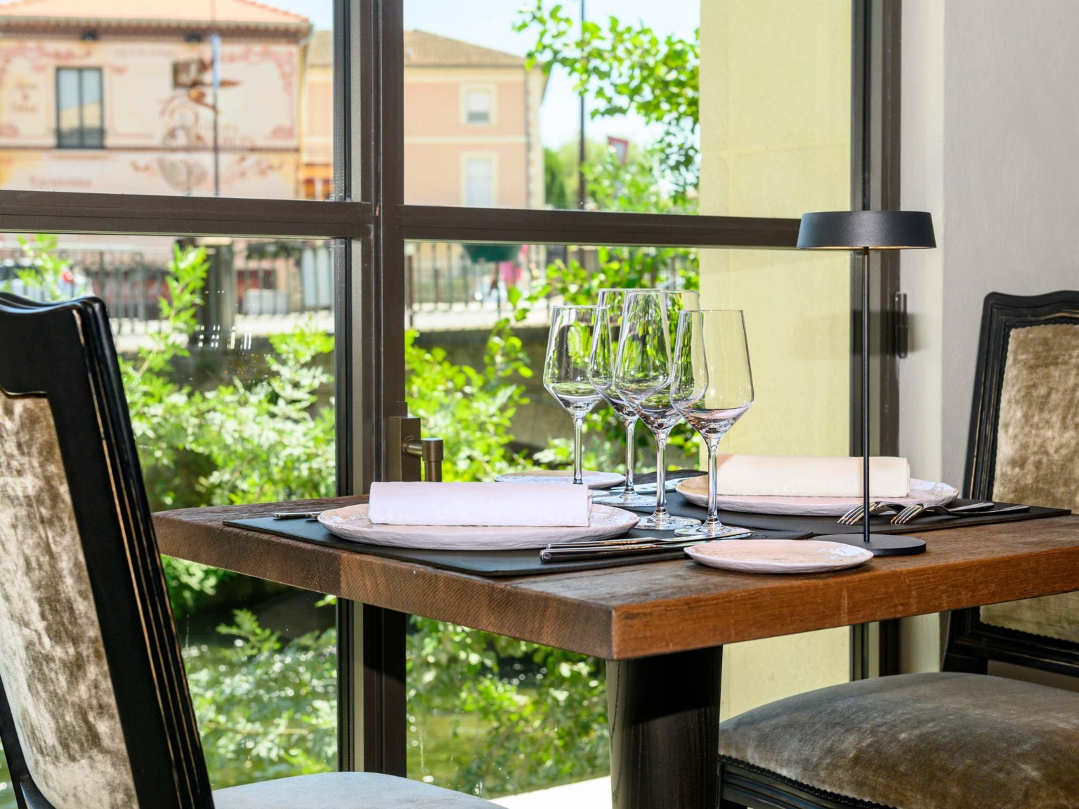 Une table de restaurant ensoleillée, dressée pour deux personnes avec des verres à vin, des assiettes et des serviettes, près d'une grande fenêtre au Dolce by Wyndham Versailles, avec vue sur la verdure et les bâtiments aux couleurs pastel du Domaine du Montcel à l'extérieur.