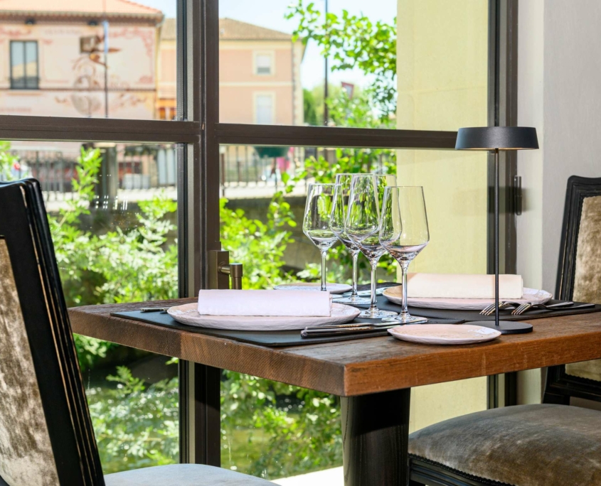 Une table de restaurant ensoleillée, dressée pour deux personnes avec des verres à vin, des assiettes et des serviettes, près d'une grande fenêtre au Dolce by Wyndham Versailles, avec vue sur la verdure et les bâtiments aux couleurs pastel du Domaine du Montcel à l'extérieur.