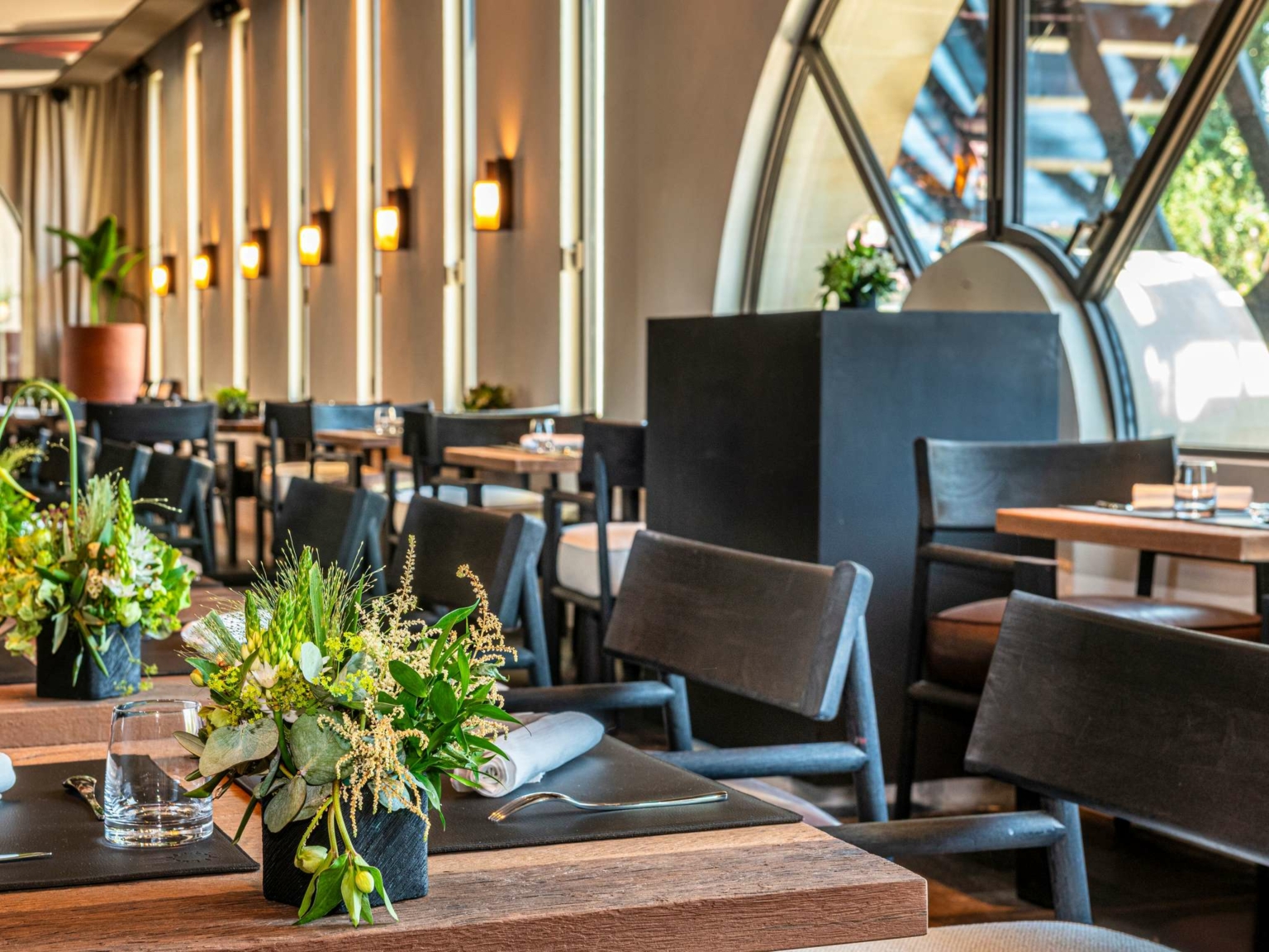 La salle à manger moderne du Dolce by Wyndham Versailles, Domaine du Montcel, se compose de tables en bois avec des centres de table en fleurs vertes, de chaises noires et d'un éclairage mural doux. De grandes fenêtres cintrées laissent entrer la lumière naturelle et offrent une vue sur la verdure luxuriante à l'extérieur.