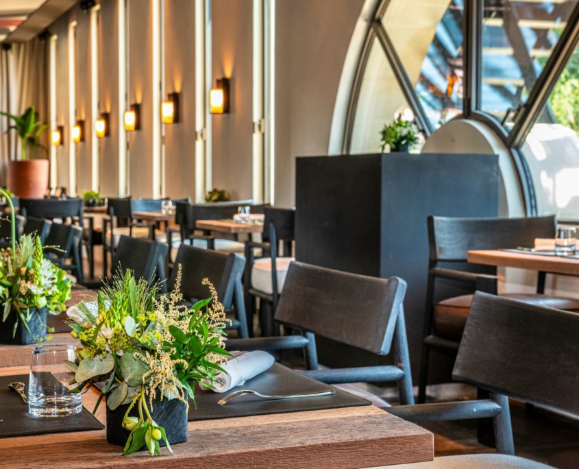 La salle à manger moderne du Dolce by Wyndham Versailles, Domaine du Montcel, se compose de tables en bois avec des centres de table en fleurs vertes, de chaises noires et d'un éclairage mural doux. De grandes fenêtres cintrées laissent entrer la lumière naturelle et offrent une vue sur la verdure luxuriante à l'extérieur.