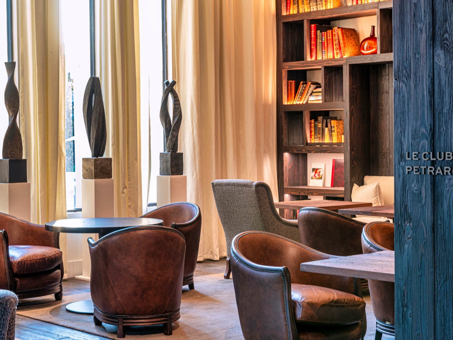 Le salon confortable du Dolce by Wyndham Versailles - Domaine du Montcel comprend des chaises en cuir marron, des tables rondes, des sculptures sur piédestal, de grandes fenêtres avec des rideaux et une étagère de livres et de décorations. Un éclairage chaleureux crée une atmosphère accueillante.