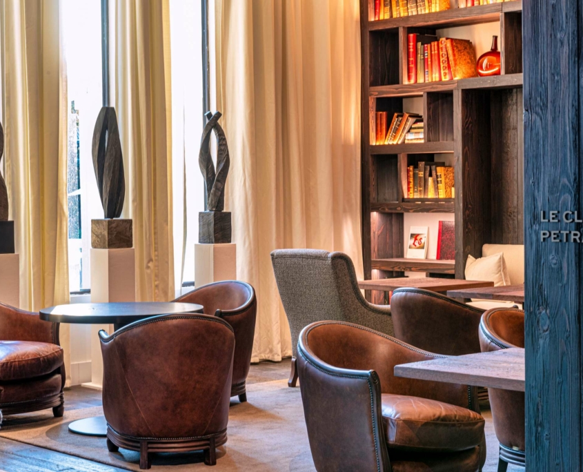 Le salon confortable du Dolce by Wyndham Versailles - Domaine du Montcel comprend des chaises en cuir marron, des tables rondes, des sculptures sur piédestal, de grandes fenêtres avec des rideaux et une étagère de livres et de décorations. Un éclairage chaleureux crée une atmosphère accueillante.