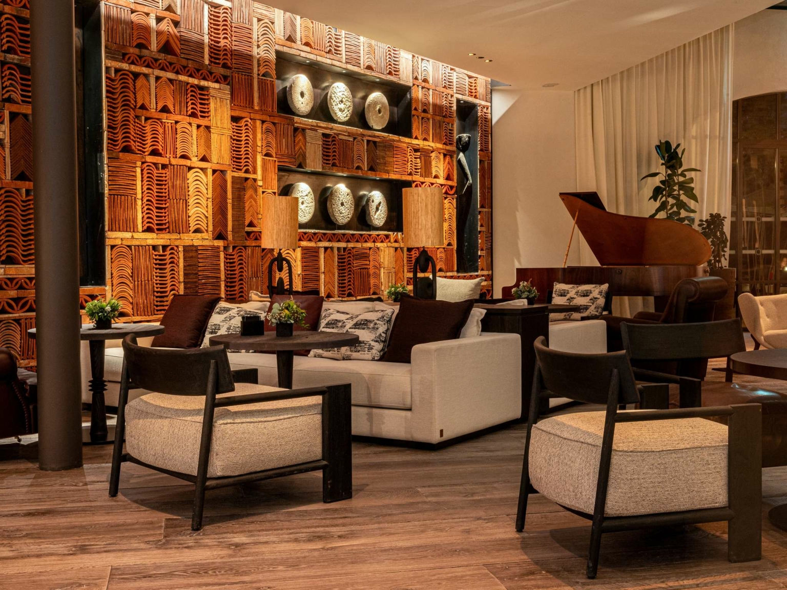 Un salon élégant au Dolce by Wyndham Versailles | Domaine du Montcel, avec des chaises et des canapés modernes, un sol en bois, des assiettes en poterie décoratives sur un mur à motifs, un éclairage chaleureux, des lampes de table, des plantes et un piano à queue en arrière-plan.