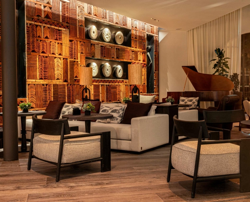 Un salon élégant au Dolce by Wyndham Versailles | Domaine du Montcel, avec des chaises et des canapés modernes, un sol en bois, des assiettes en poterie décoratives sur un mur à motifs, un éclairage chaleureux, des lampes de table, des plantes et un piano à queue en arrière-plan.