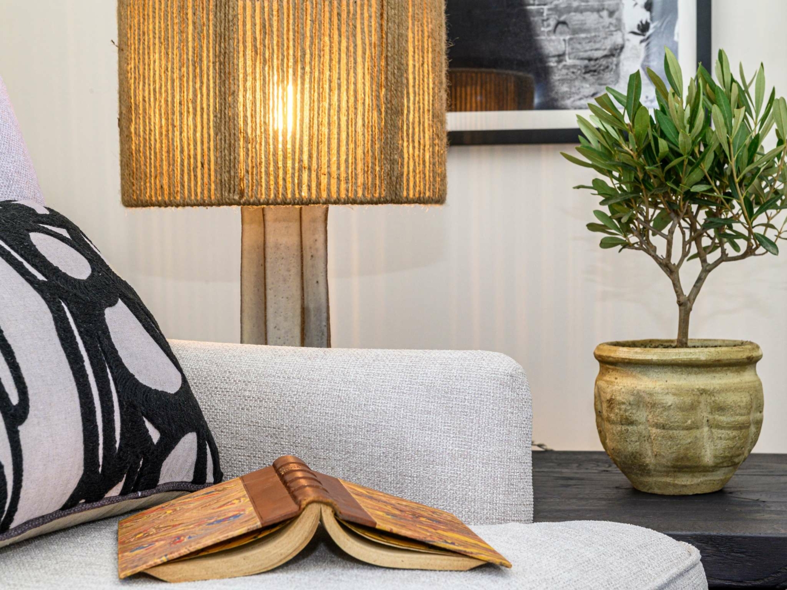 Une scène de salon confortable inspirée de Dolce by Wyndham Versailles, avec un fauteuil beige, un coussin noir et blanc, un livre ouvert, une lampe de table allumée avec un abat-jour texturé, et une plante en pot sur une table sombre à côté du fauteuil.
