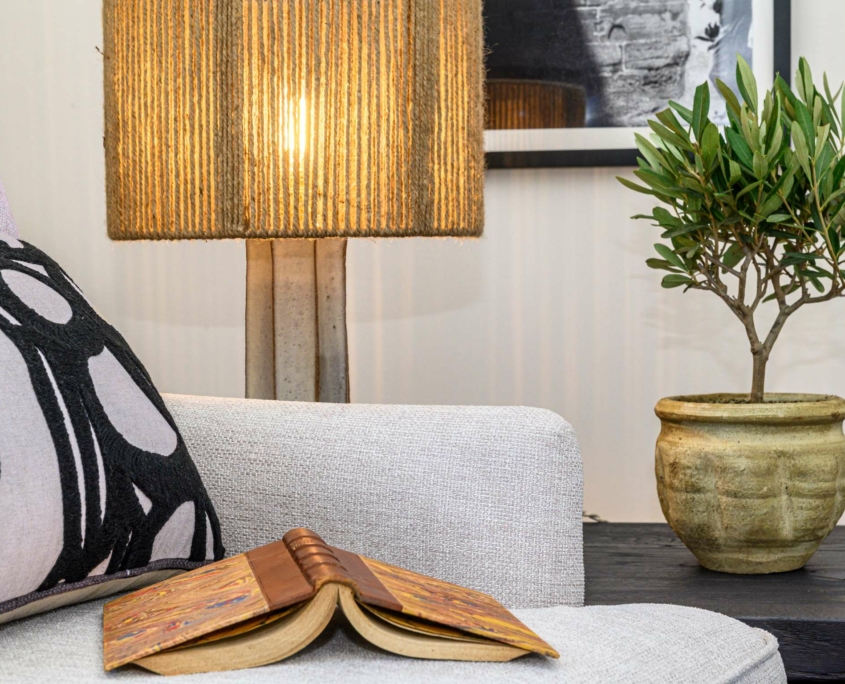 Une scène de salon confortable inspirée de Dolce by Wyndham Versailles, avec un fauteuil beige, un coussin noir et blanc, un livre ouvert, une lampe de table allumée avec un abat-jour texturé, et une plante en pot sur une table sombre à côté du fauteuil.