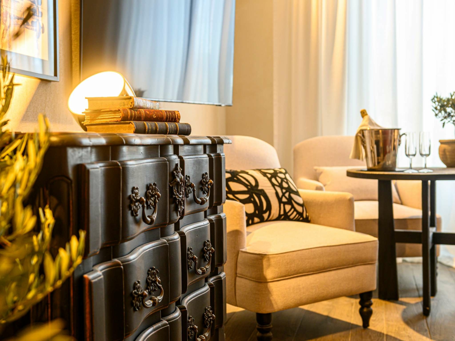 Le salon confortable du Dolce by Wyndham Versailles | Domaine du Montcel est composé de fauteuils crème, d'une table ronde avec un seau à champagne et des verres, d'une commode sombre ornée de livres et d'une lampe, le tout baigné d'une douce lumière naturelle grâce à de grands rideaux.