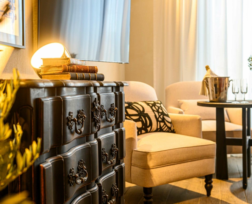 Le salon confortable du Dolce by Wyndham Versailles | Domaine du Montcel est composé de fauteuils crème, d'une table ronde avec un seau à champagne et des verres, d'une commode sombre ornée de livres et d'une lampe, le tout baigné d'une douce lumière naturelle grâce à de grands rideaux.