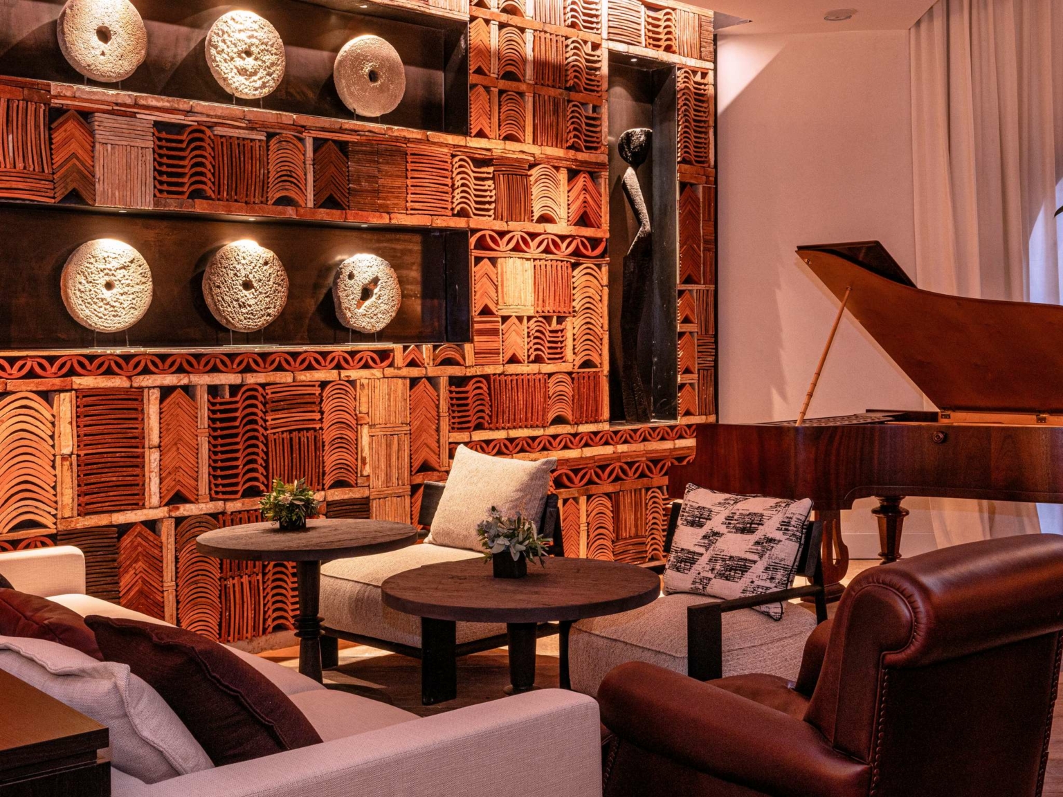 Le salon confortable du Dolce by Wyndham Versailles | Domaine du Montcel comprend des fauteuils modernes, une chaise en cuir, une table basse ronde, un piano à queue et un mur texturé orné de motifs géométriques, d'œuvres d'art sculptées et d'un éclairage doux.
