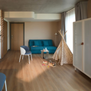 Une chambre d'hôtel moderne avec un sol en bois, un canapé bleu, une chaise blanche, une petite table et une tente tipi ludique affiche Agencement sur-mesure : la nouvelle intelligence du design en 2026 - la lumière remplit l'espace à travers de grandes fenêtres aux rideaux gris.
