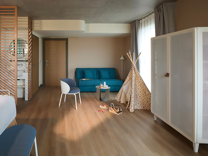 Une chambre d'hôtel moderne avec un sol en bois, un canapé bleu, une chaise blanche, une petite table et une tente tipi ludique affiche Agencement sur-mesure : la nouvelle intelligence du design en 2026 - la lumière remplit l'espace à travers de grandes fenêtres aux rideaux gris.