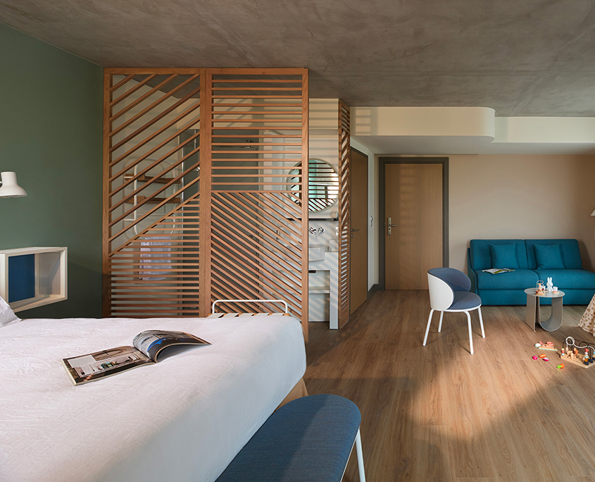 Chambre moderne de l'hôtel Okko Troyes avec un sol en bois, un lit sur lequel est posé un magazine, un canapé bleu, une chaise, des jouets éparpillés et une cloison en lattes de bois qui sépare la chambre de la salle de bains.