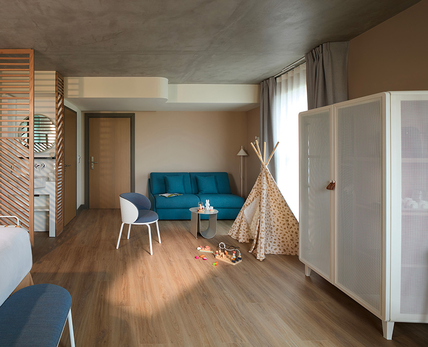 La chambre spacieuse et moderne de l'hôtel Okko Troyes est dotée d'un sol en bois, d'un canapé bleu, d'une chaise blanche et bleue, d'une armoire blanche et d'une tente de jeu beige. Des jouets sont éparpillés près de la tente, tandis qu'une lumière douce filtre à travers des rideaux diaphanes.