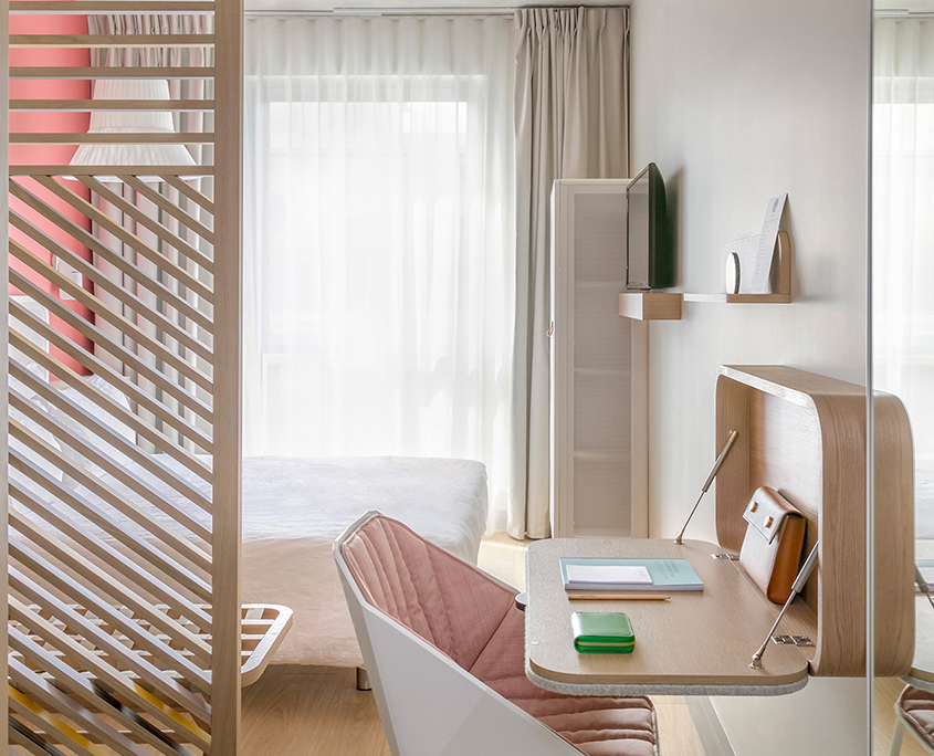Chambre et espace de travail minimalistes à l'hôtel Okko de Troyes, baignés de lumière naturelle. Elle comprend un bureau en bois, une chaise rose rembourrée, des étagères flottantes et une cloison en lattes de bois qui sépare le lit de l'élégant espace de travail.