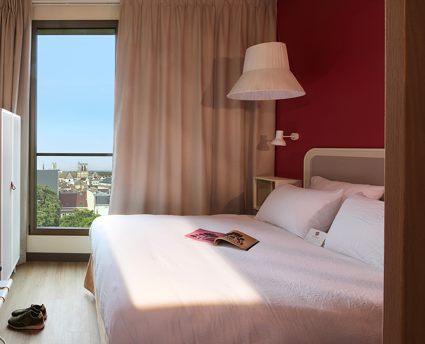 À l'hôtel Okko de Troyes, un lit soigneusement fait avec des draps blancs se trouve près d'une fenêtre aux rideaux beiges, avec vue sur un paysage urbain. Un magazine ouvert est posé sur le lit et une paire de chaussures est posée sur le sol, près du pied du lit.