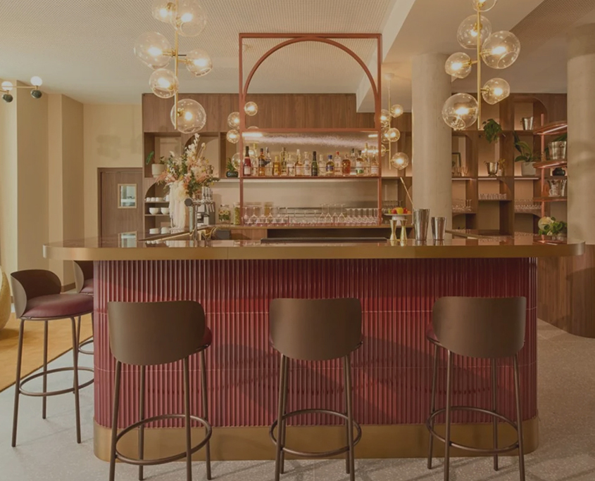Le bar moderne de l'hôtel Okko Troyes se compose d'un comptoir rouge, d'éléments en laiton, de quatre tabourets sombres, de lampes suspendues, d'étagères contenant des bouteilles et des verres, et de compositions florales dans une pièce spacieuse et bien éclairée.