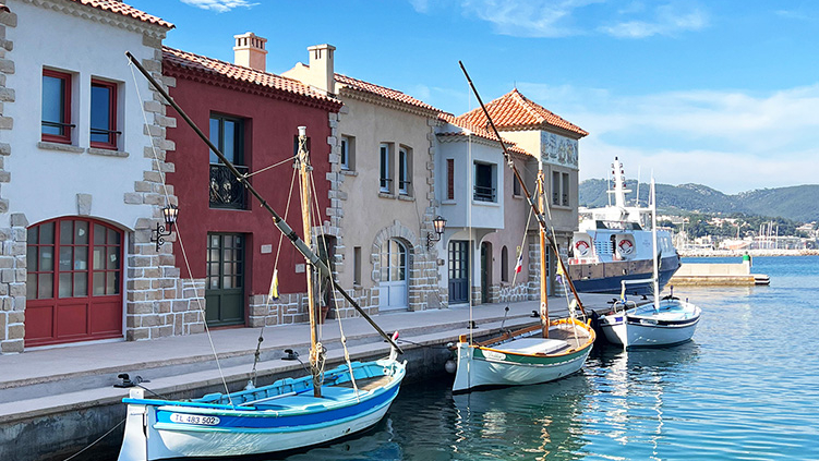 Des bateaux de pêche aux couleurs vives sont amarrés le long d'un front de mer bordé de charmants bâtiments de style méditerranéen, sous un ciel bleu. L'eau limpide reflète les bateaux et les bâtiments, créant un décor qui rappelle les séjours à l'Okko Hotels Troyes.